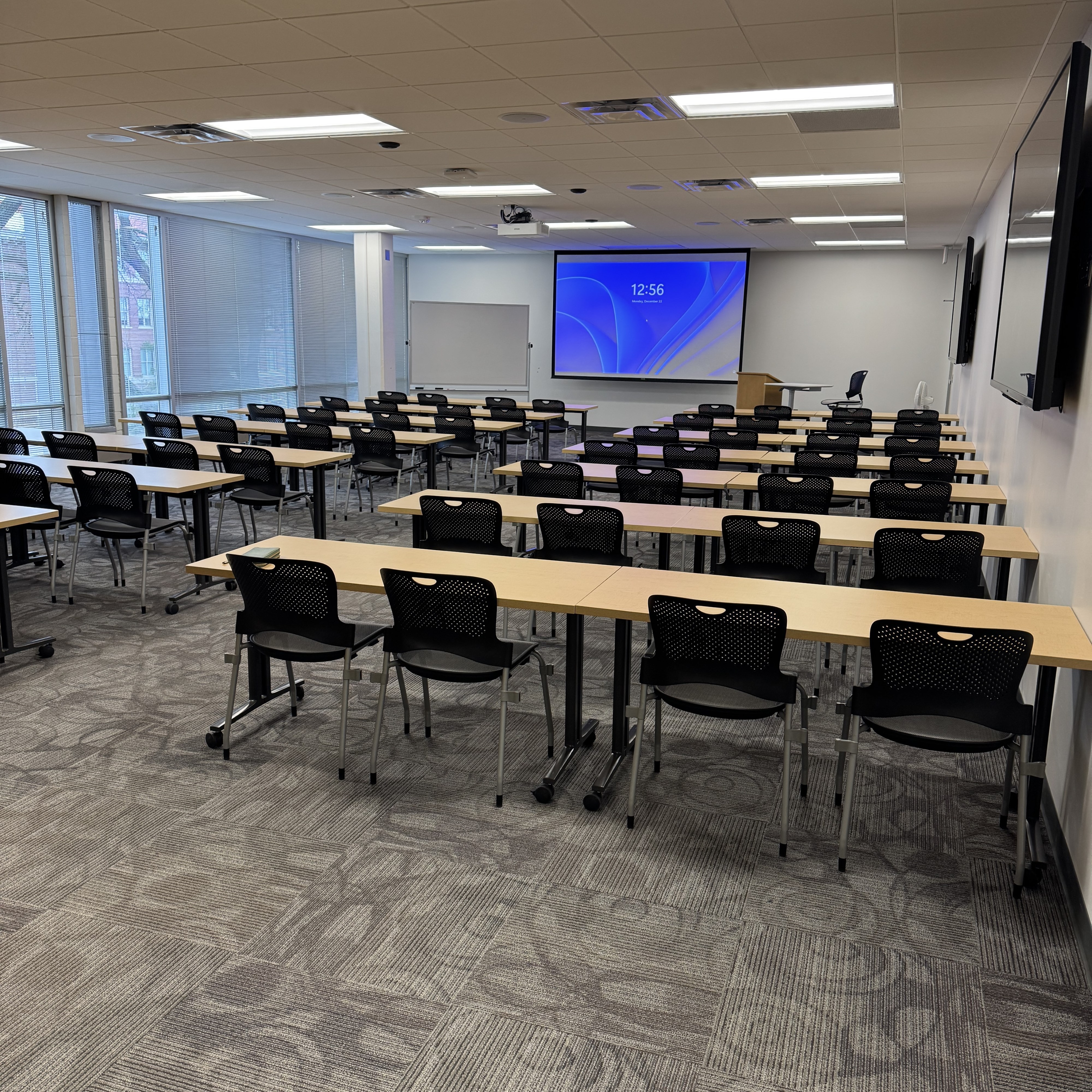 ScholarSpace room 301 in Rod Library. A projector screen and podium are located at the front of the room. The room also contains wall-mounted computer monitors, tables, and chairs.